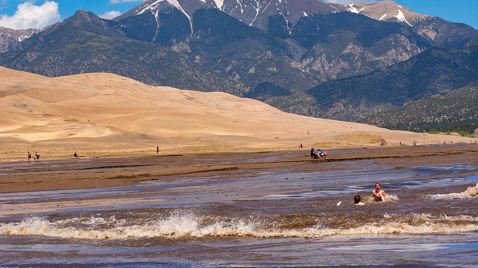 Children playing in a wave surging down a wide creek at the base of the dunes and mountains