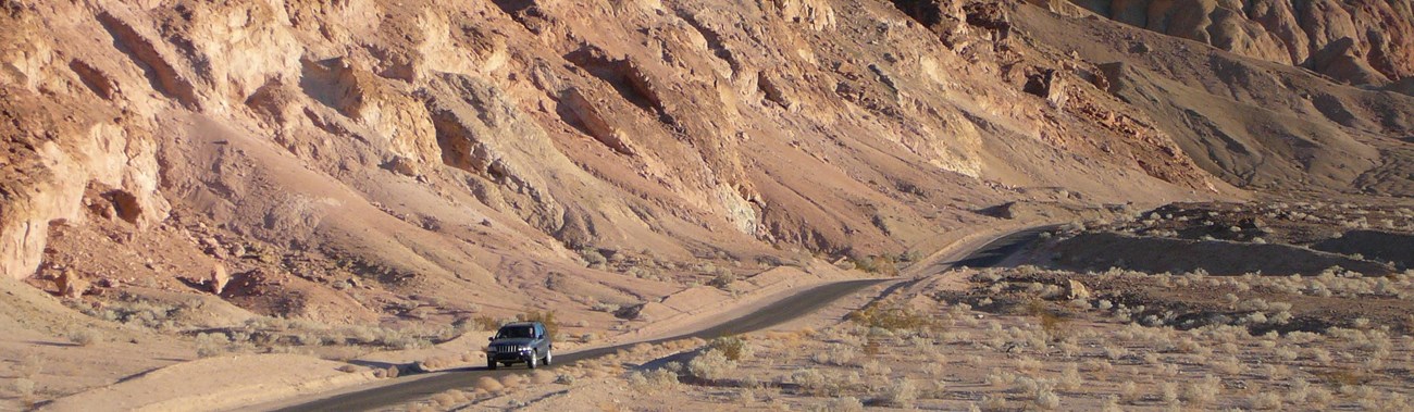 A vehicle drives along a single lane road through colorful, eroded hills. 