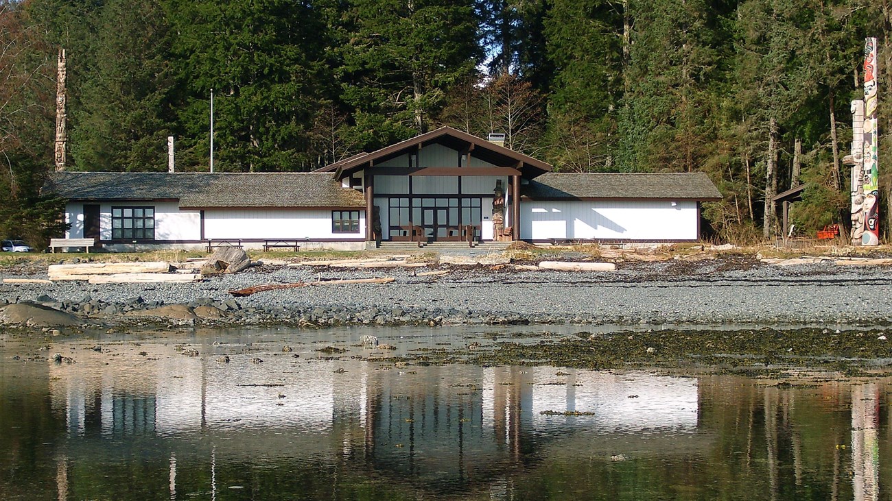 Calm water with a low, white building flanked by totem poles in the background