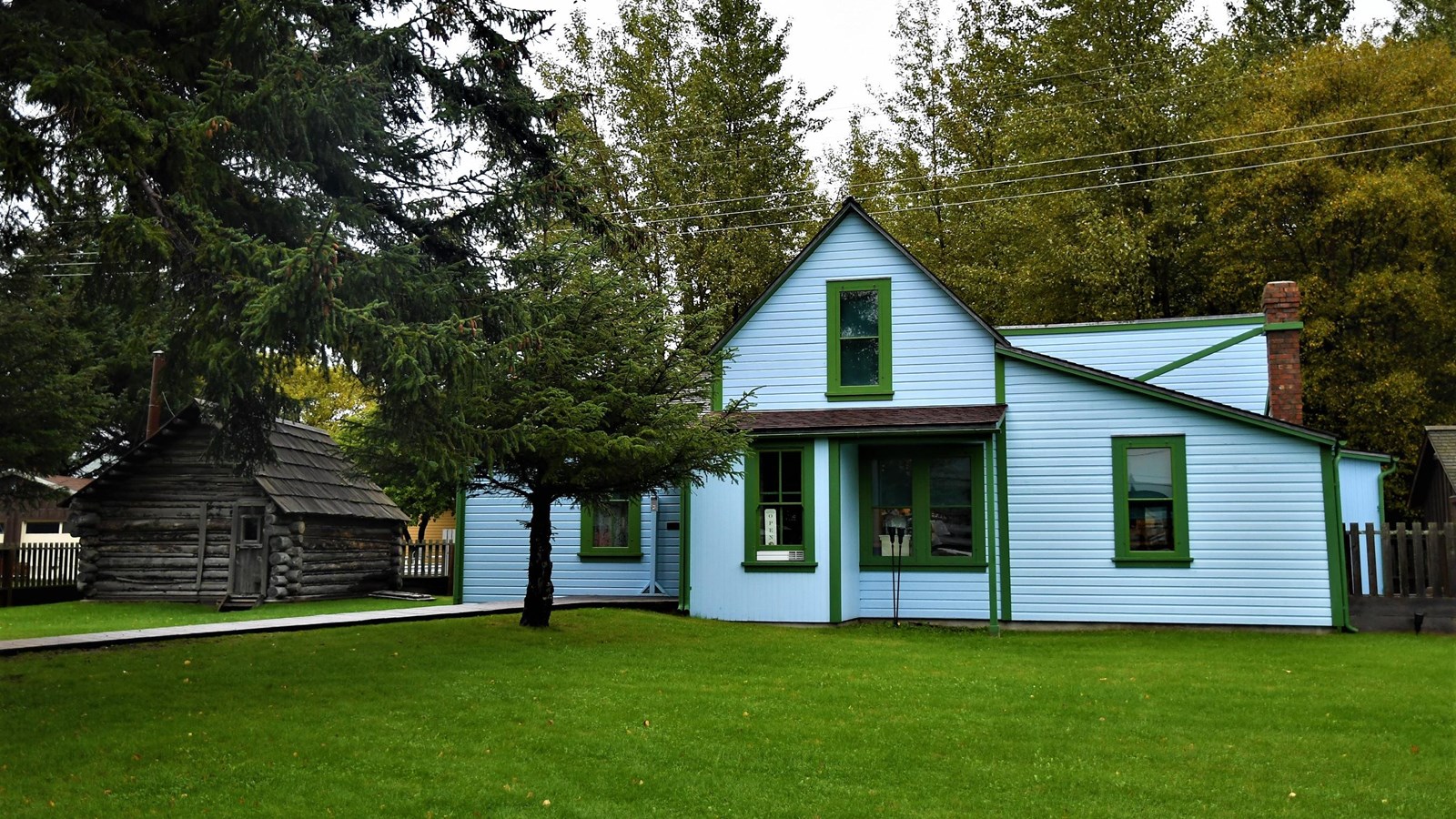 A small log cabin and a blue wood frame house with green trim surrounded by grass and trees.