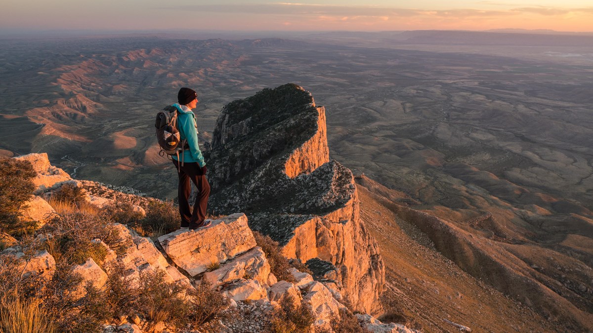 A hiker stands on a peak with lower mountains in the background at sunset