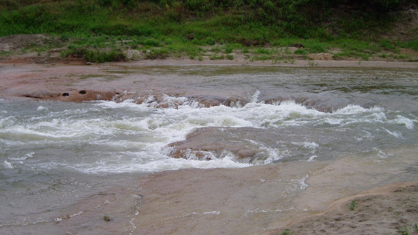 A river runs over a shelf and creates a rapid and white water with stone ledges on each side.