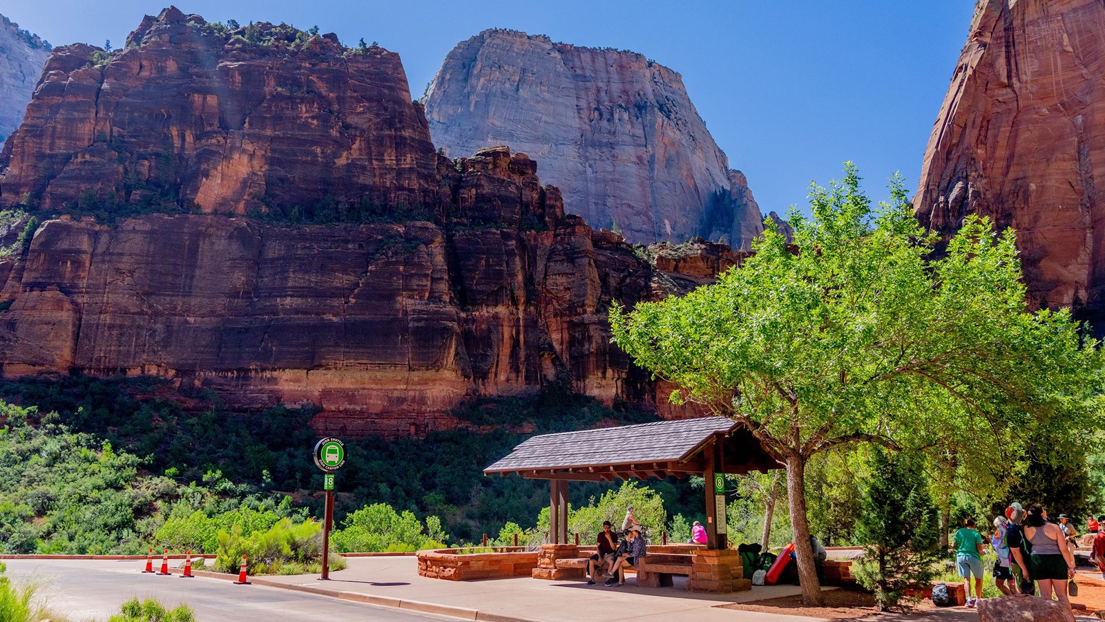 A group of visitors under a shaded shuttle stop surrounded by rocks and green foliage.