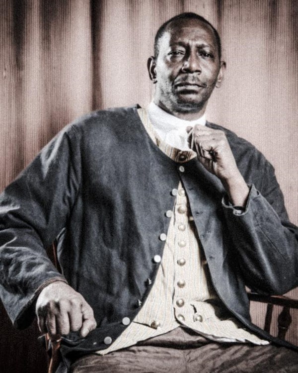 A brown man in a wool suit sits with his fist clenched in front of him in thought.