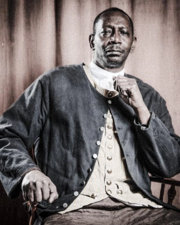 A brown man in a wool suit sits with his fist clenched in front of him in thought.