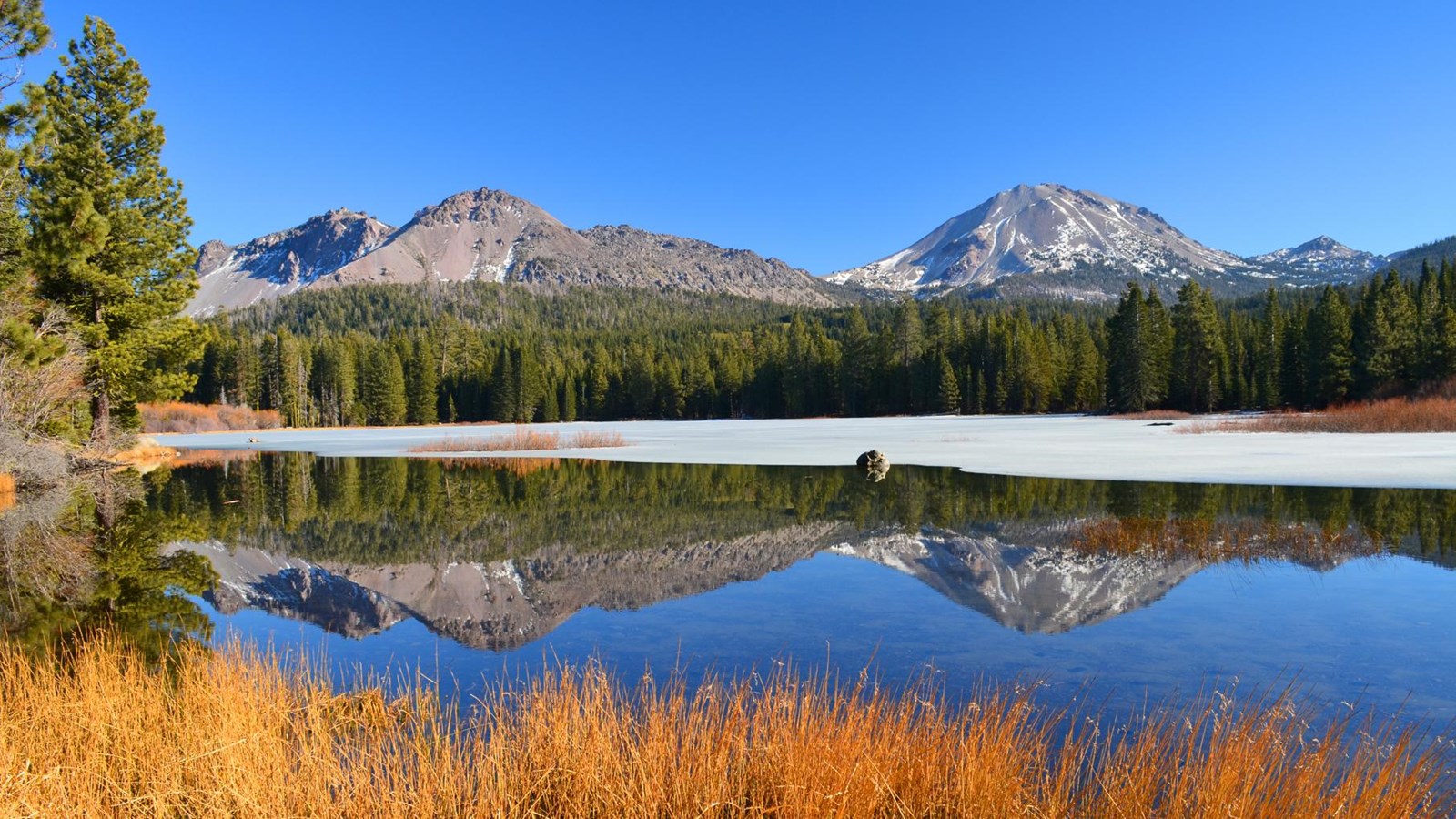 Two volcanic peaks reflected in a partially frozen lake lined by golden grass and evergreen trees. 