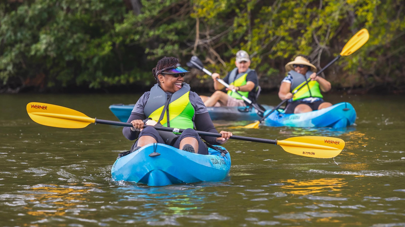 Three adults kayaking with bright green life jackets on a calm lake, surrounded by greenery.