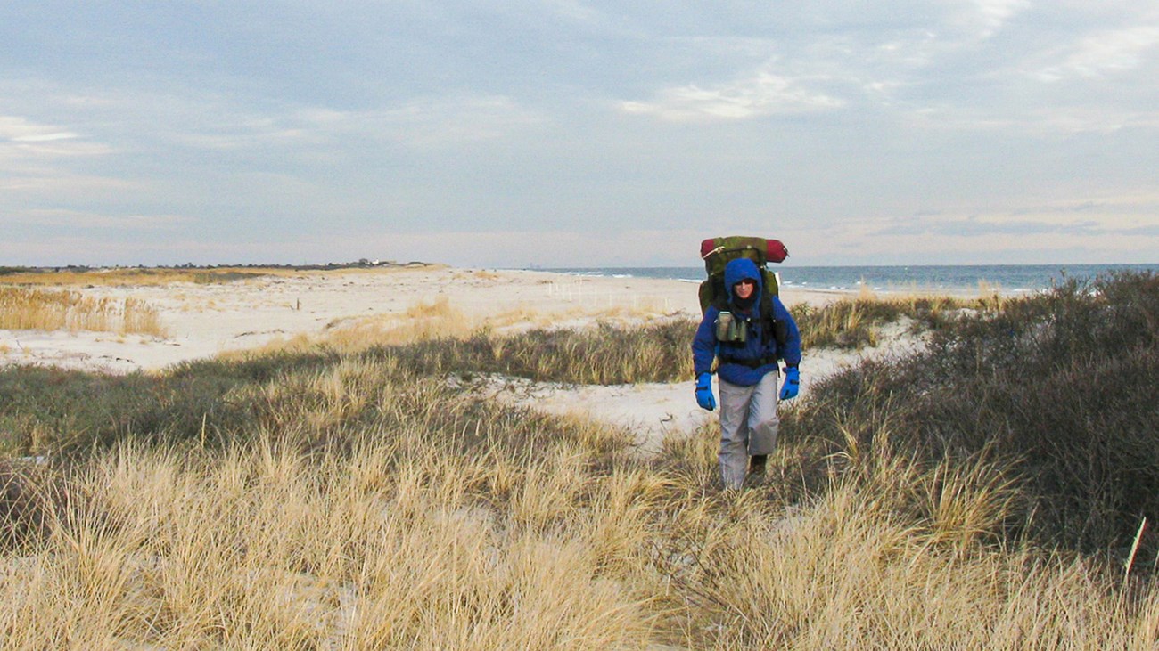 A backpacker with winter clothes walks a grassy dune near the ocean.