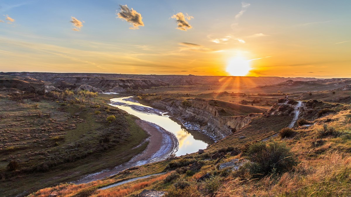 A bend in the river and a trail above it are seen at sunset. Buttes and prairies line the river.