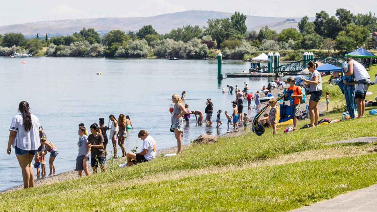 Color photograph of a river\'s shore with numerous people gathered on a green lawn beside it.