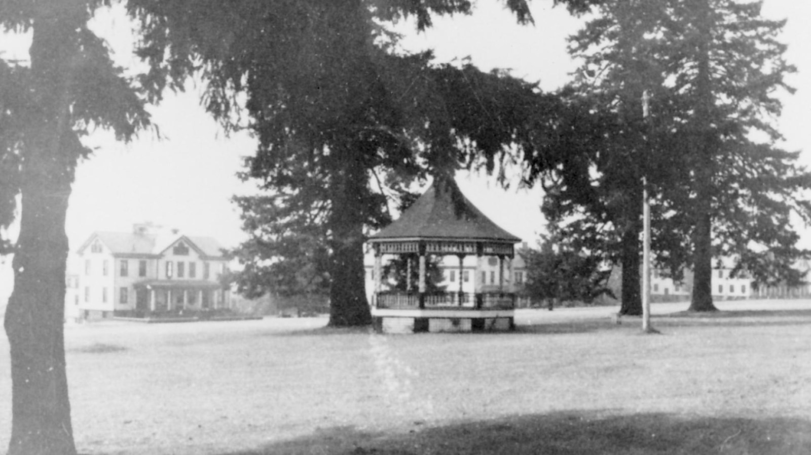 A black and white photo shows a gazebo in a grassy field surrounded by trees and buildings.