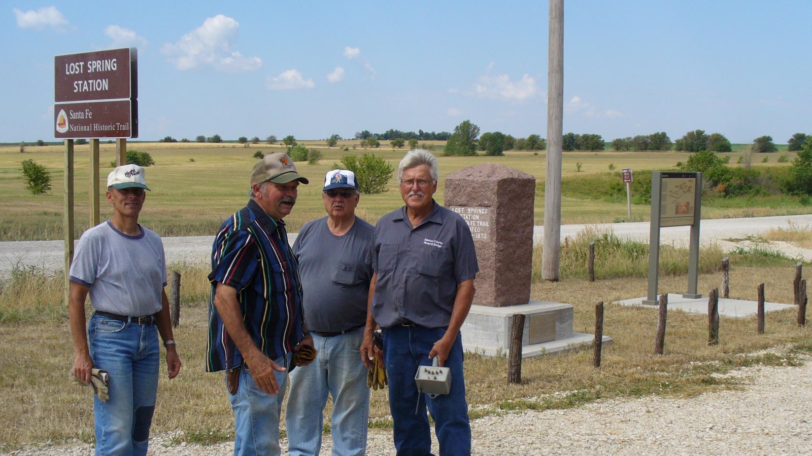 A group of men stand next to a site identification sign and a stone marker in a grassy prairie.