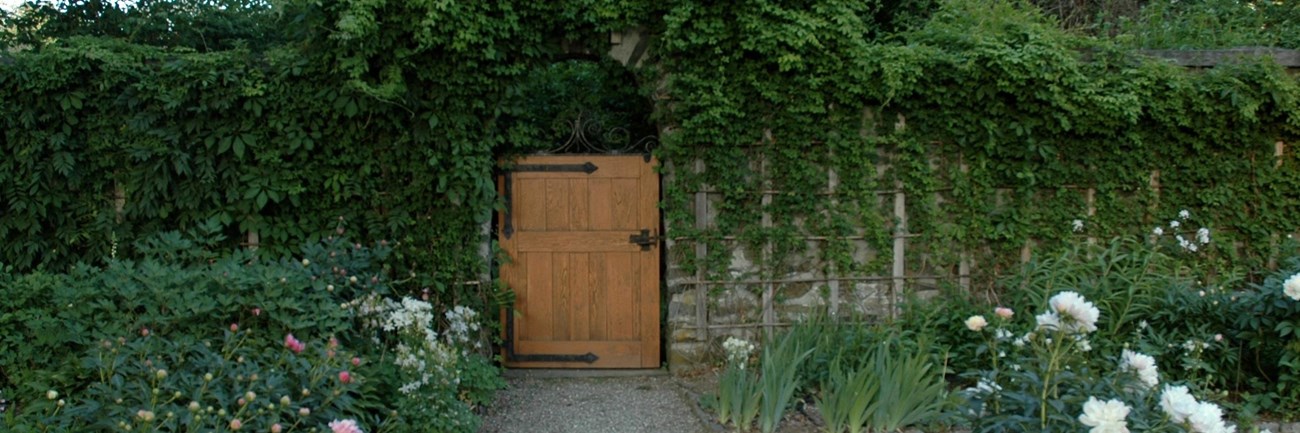 A garden gate surrounded by stone wall and flowering plants in foreground.