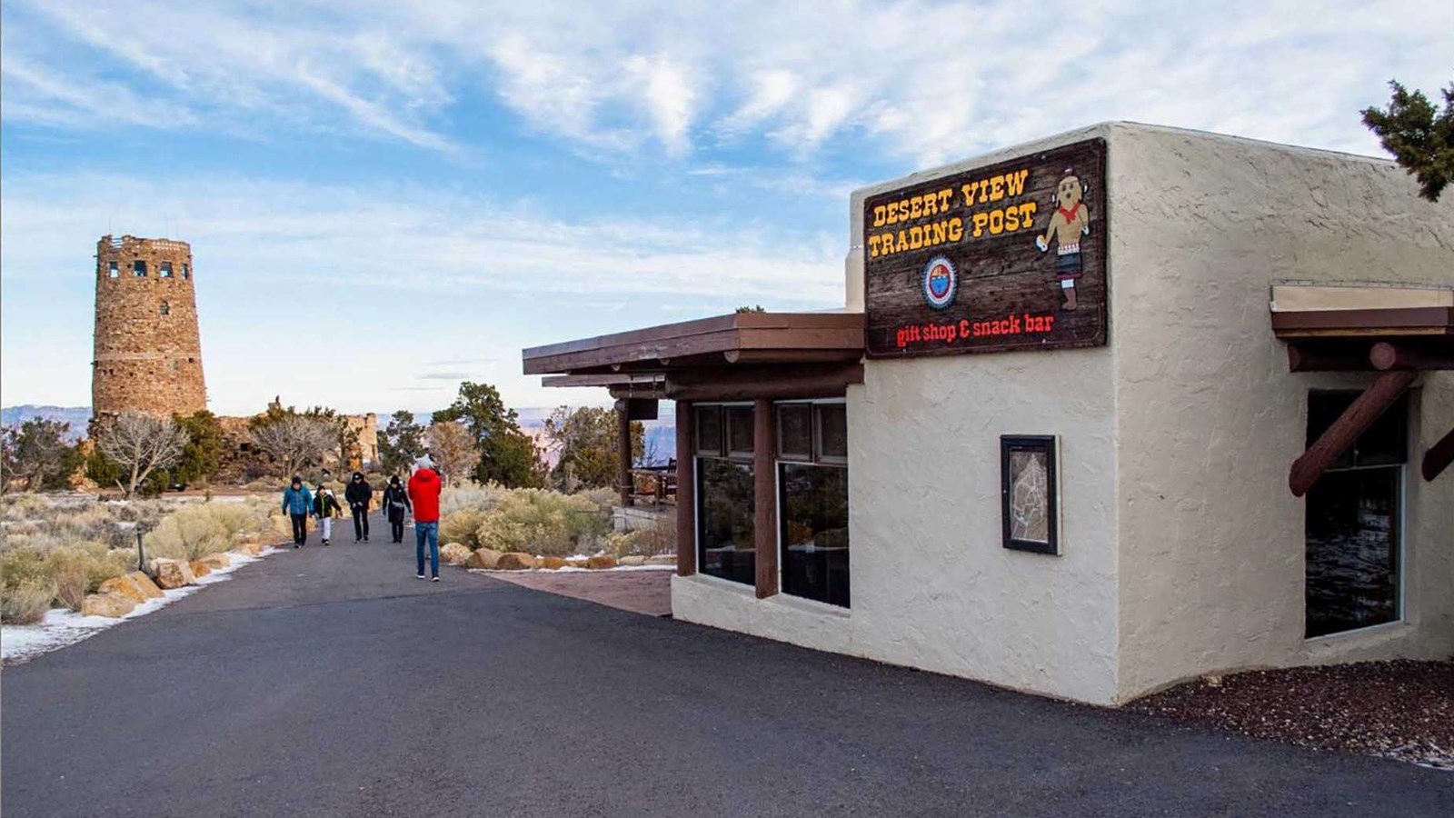 An white adobe style building sits on a paved path that leas straight to the Desert View Watchtower.