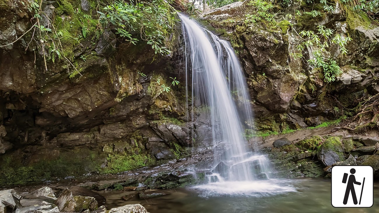A waterfall surrounded by rocks, moss, and plants. Trail leads behind it. A hiker icon in corner.