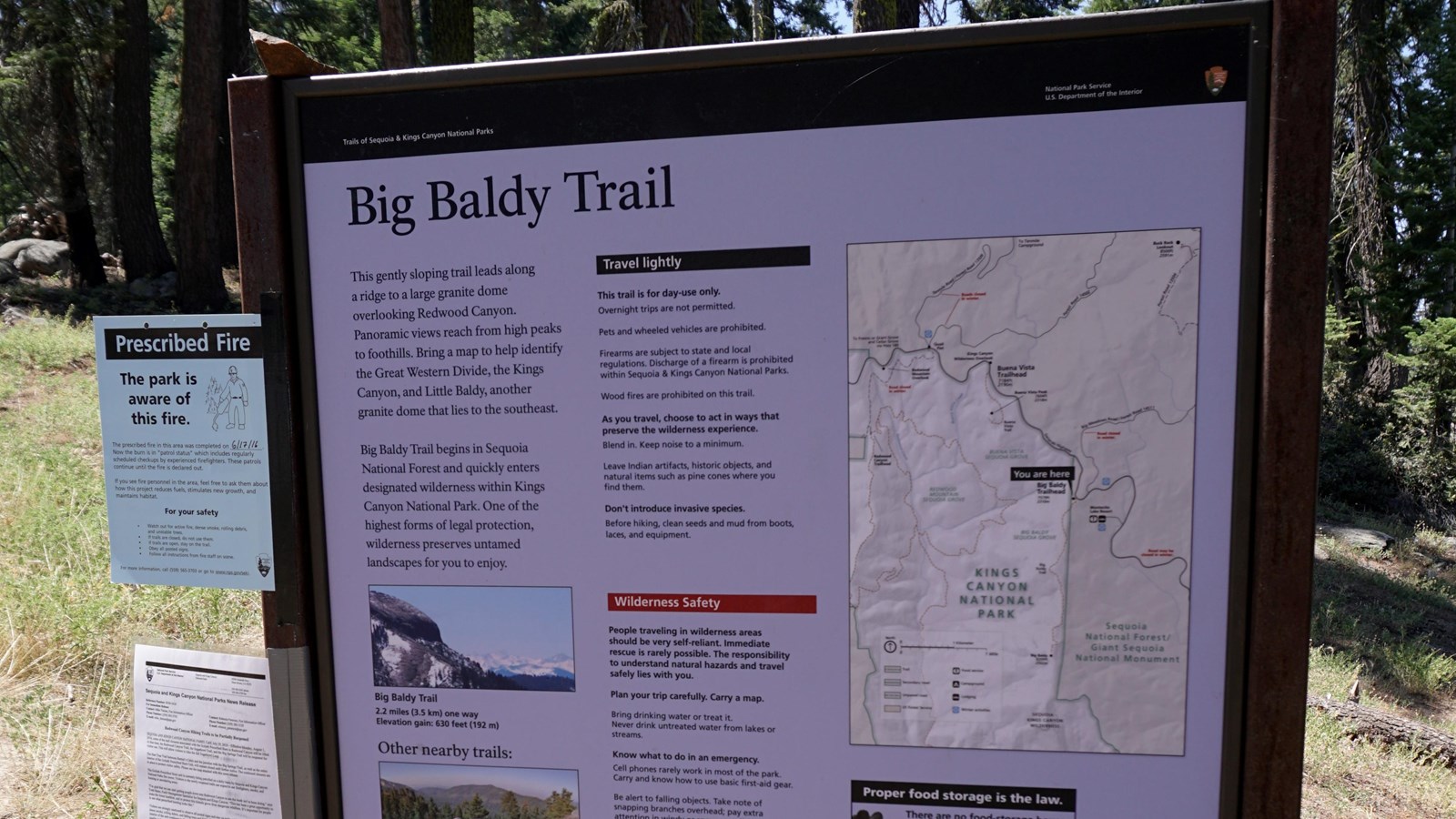 A metal framed sign sits i front a dirt path leading to a stand of trees