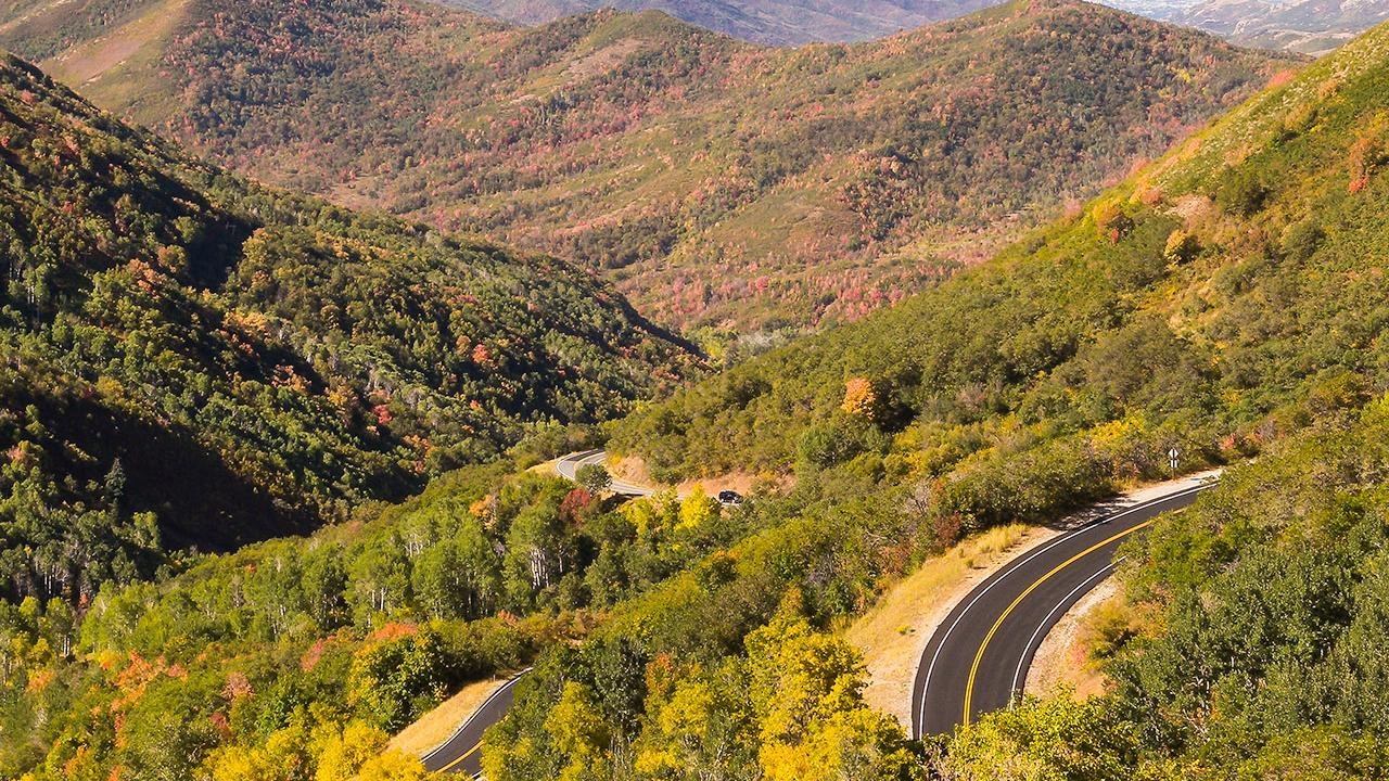 A twisted canyon road descends through hills covered with fall vegetation.