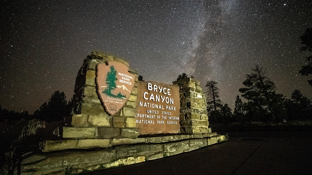 An illuminated Bryce Canyon park entrance sign against a backdrop of stars in a dark sky