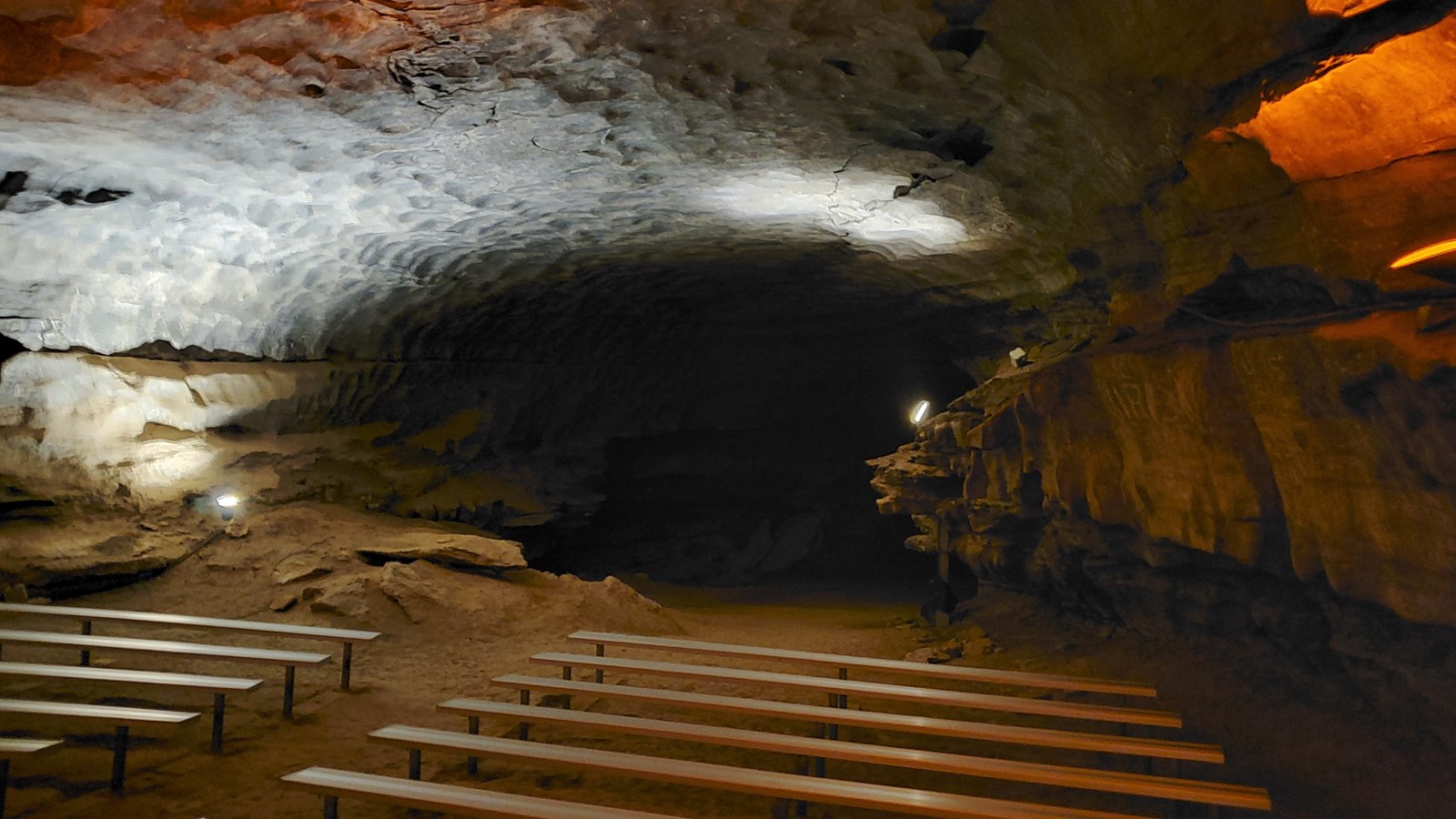 Wooden benches sit in the center of a large cave passage with a dark passage in the background. 