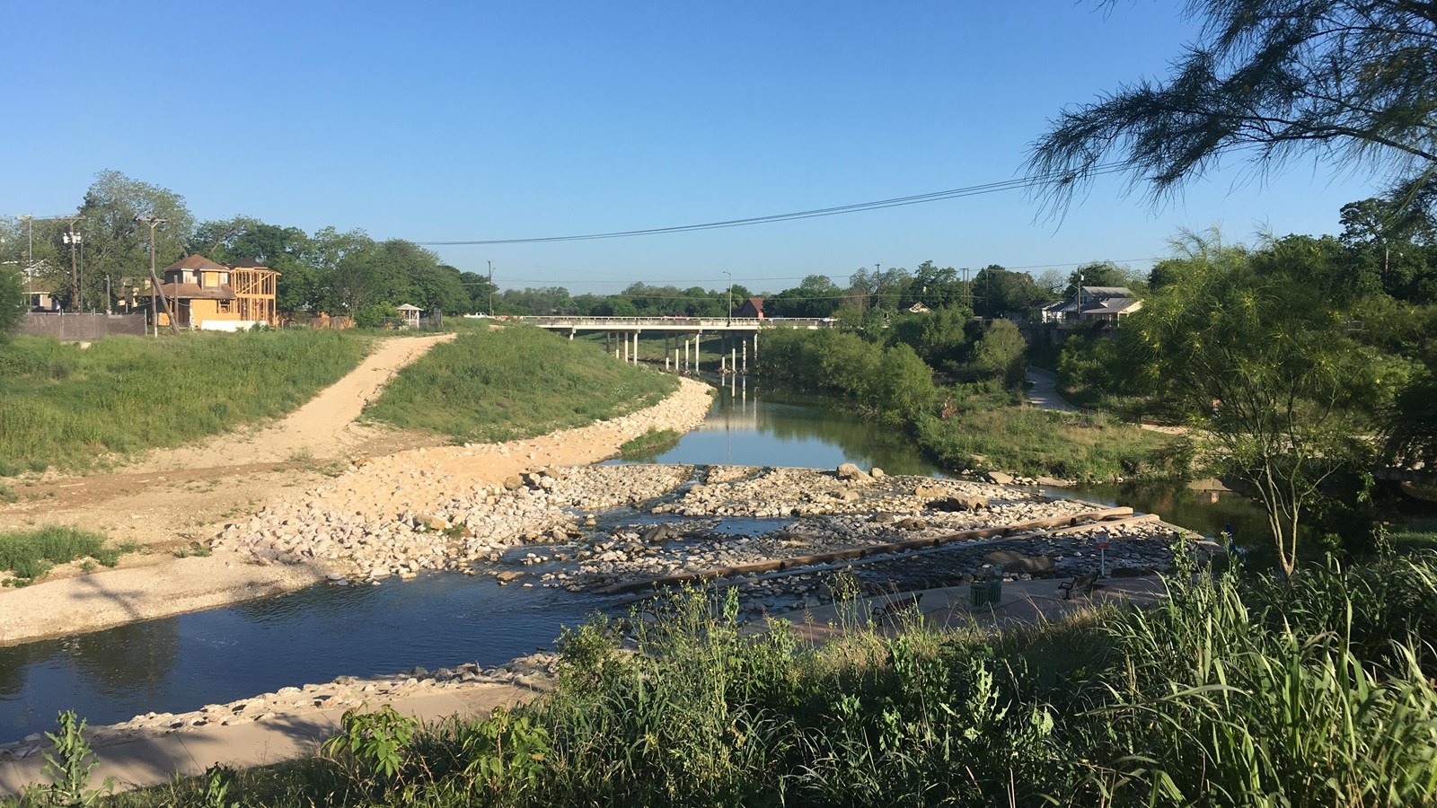 Paved trail bends along the San Antonio River with a vehicular bridge in background