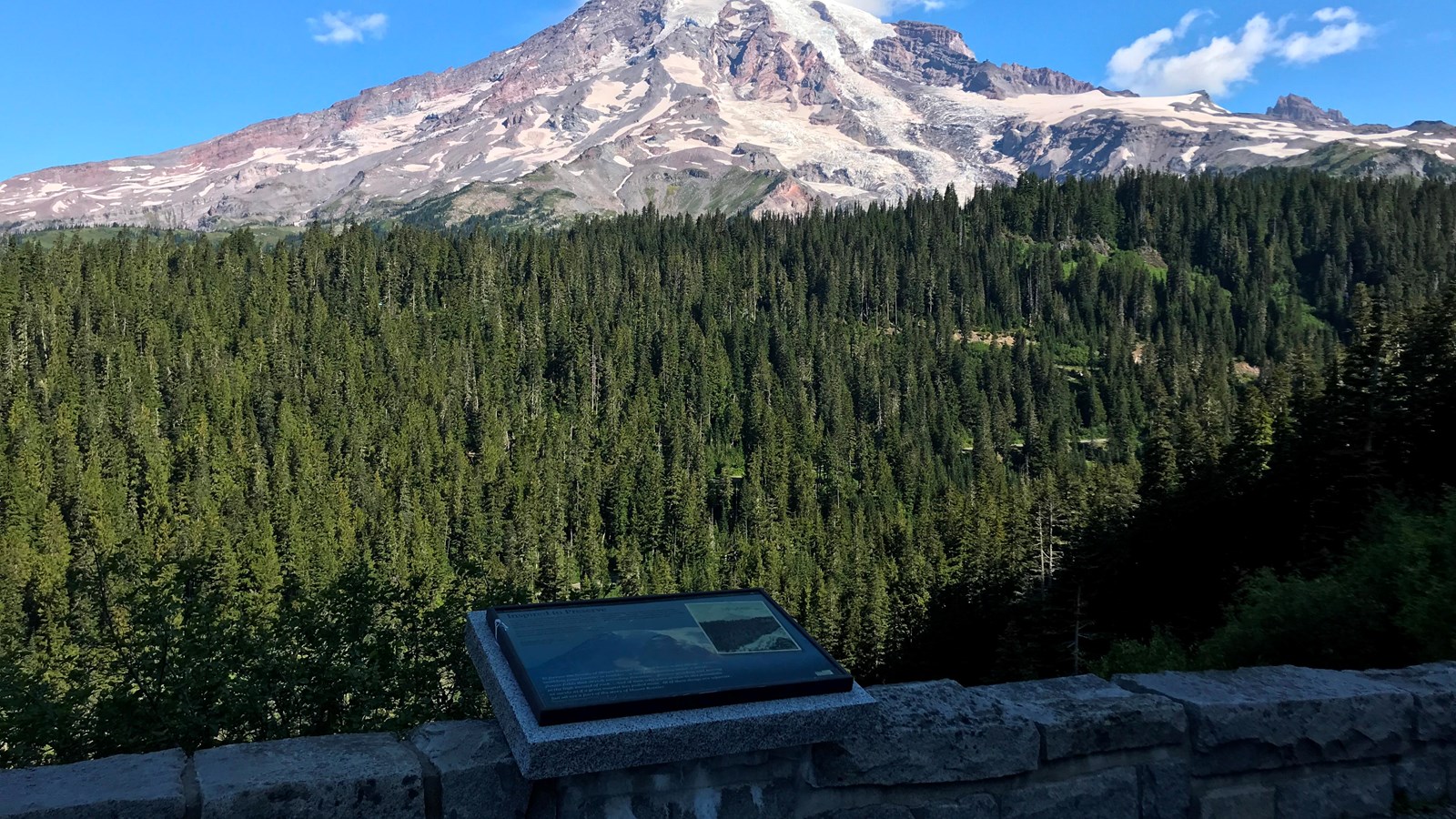 A exhibit panel on a low rock wall overlooks a forested valley and a glaciated mountain peak.