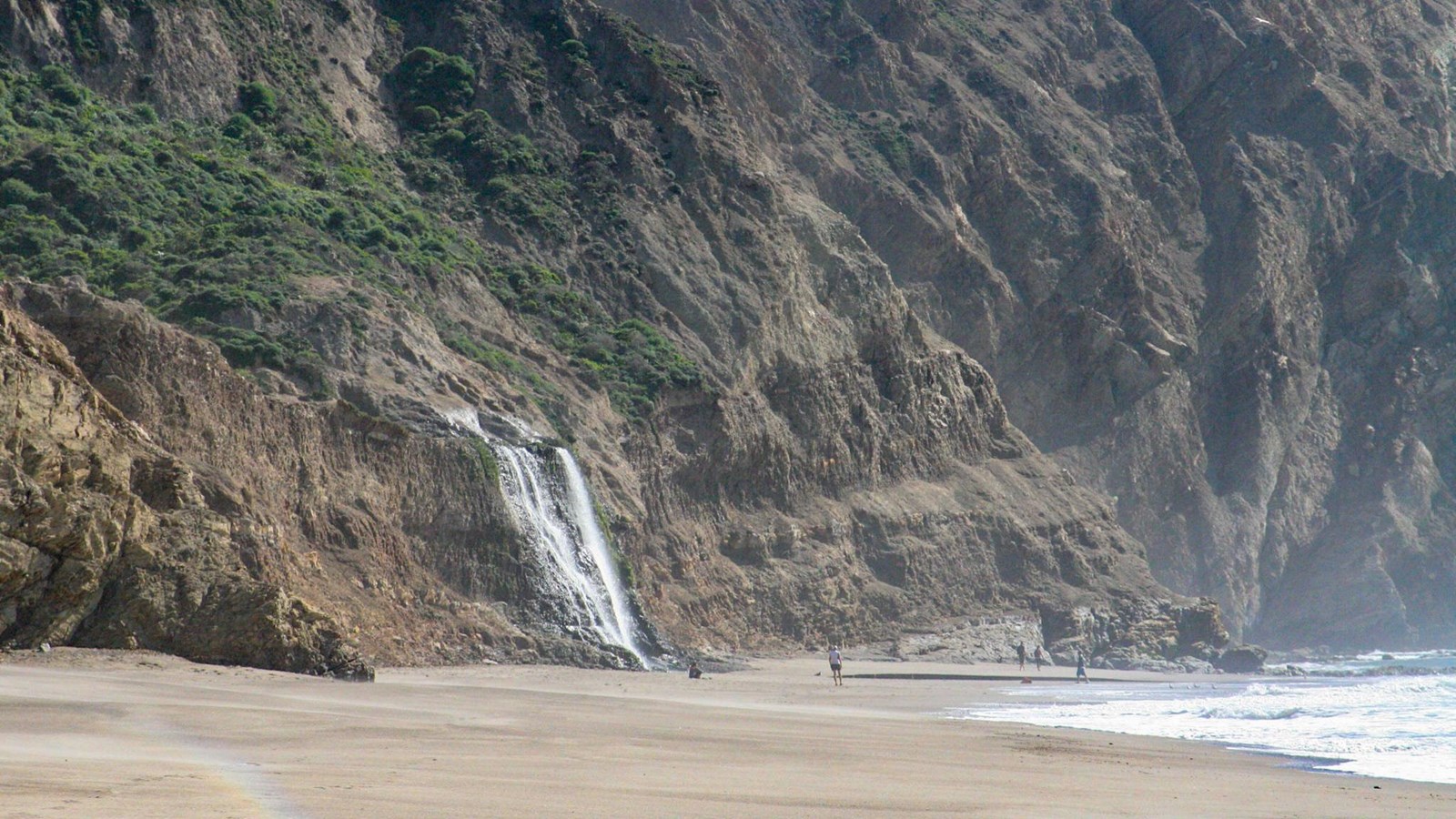 A 40-foot-tall waterfall cascades off of a coastal bluff onto a sandy ocean beach..
