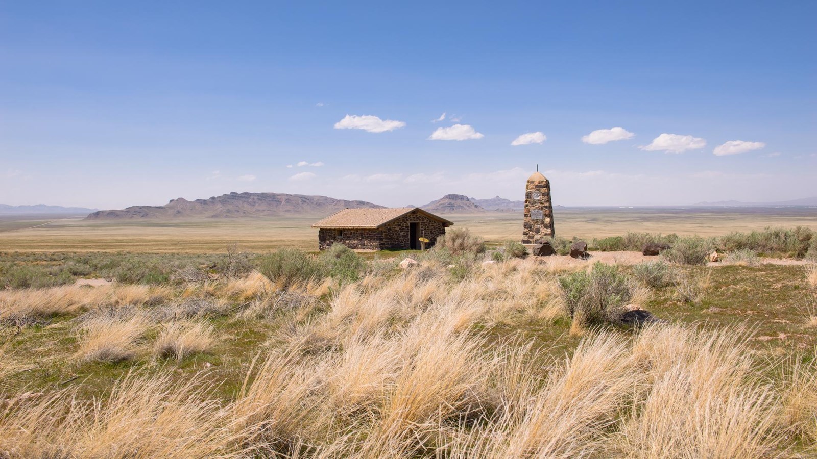 A one room, pitched roof stone building sits in a vast open flat area, next to a stone monument.