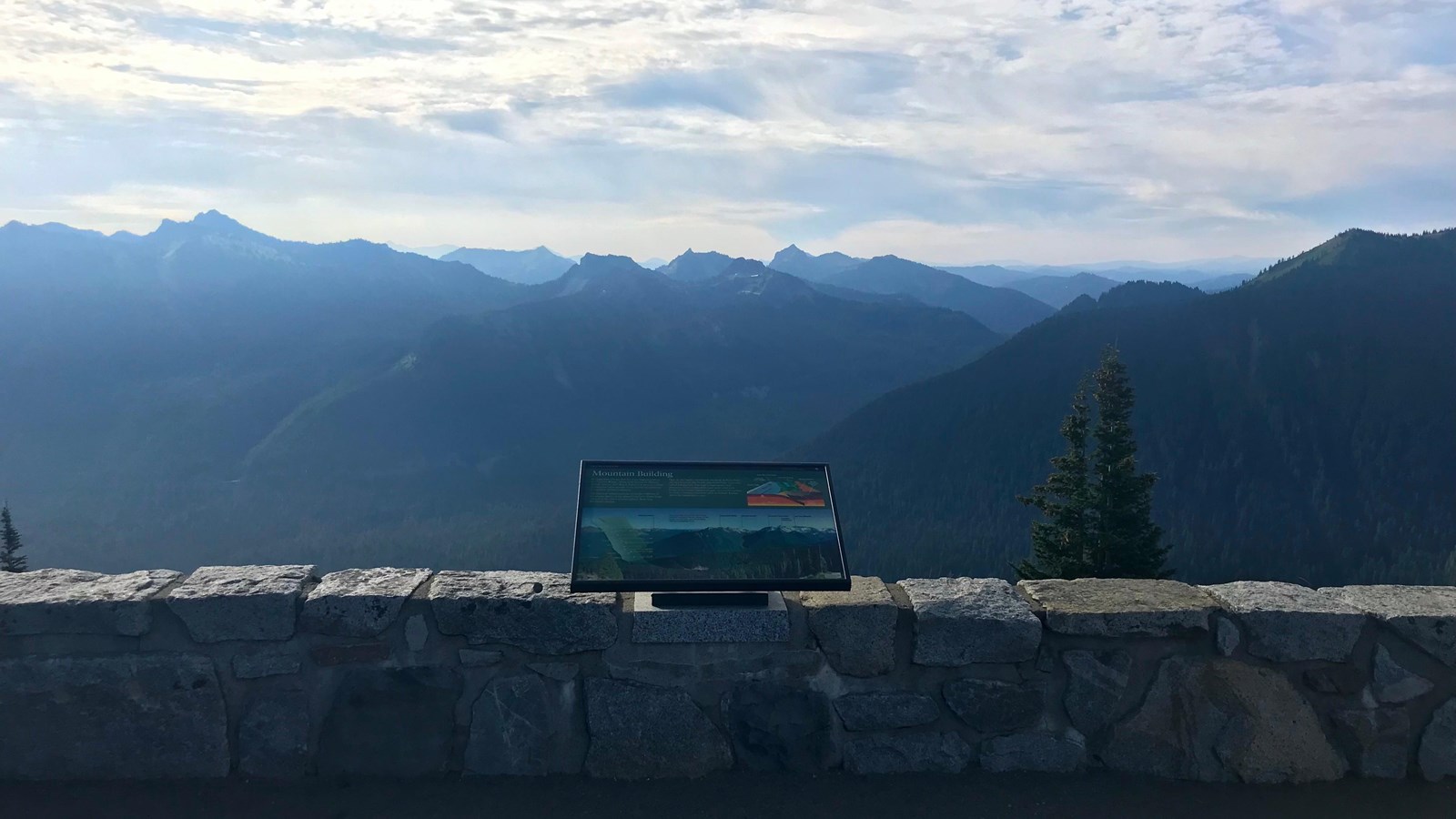 A wayside panel attached to a rock wall overlooks a mountain range and deep valleys.