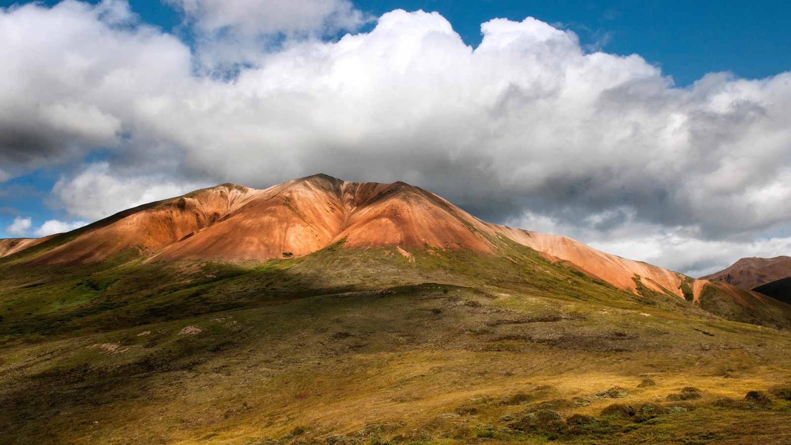 Alpine tundra and rust colored rocks on a mountainside against a blue sky with fluffy white clouds
