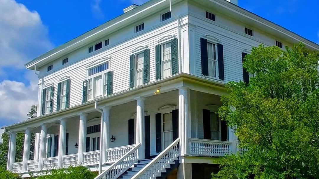 A white two-story house has green shutters and is surrounded by greenery.