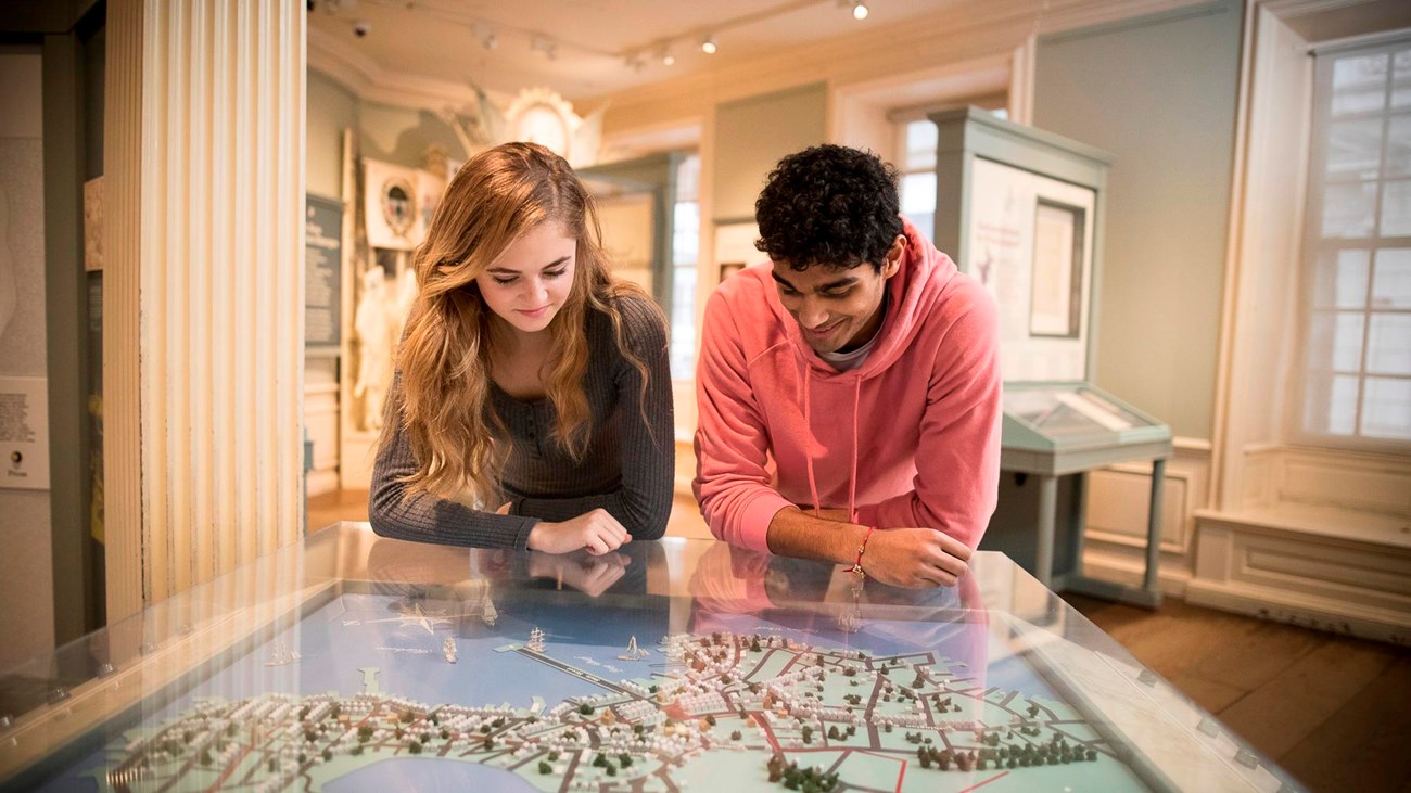Two youth in an exhibit space, leaning over an interactive map table and looking down at it.