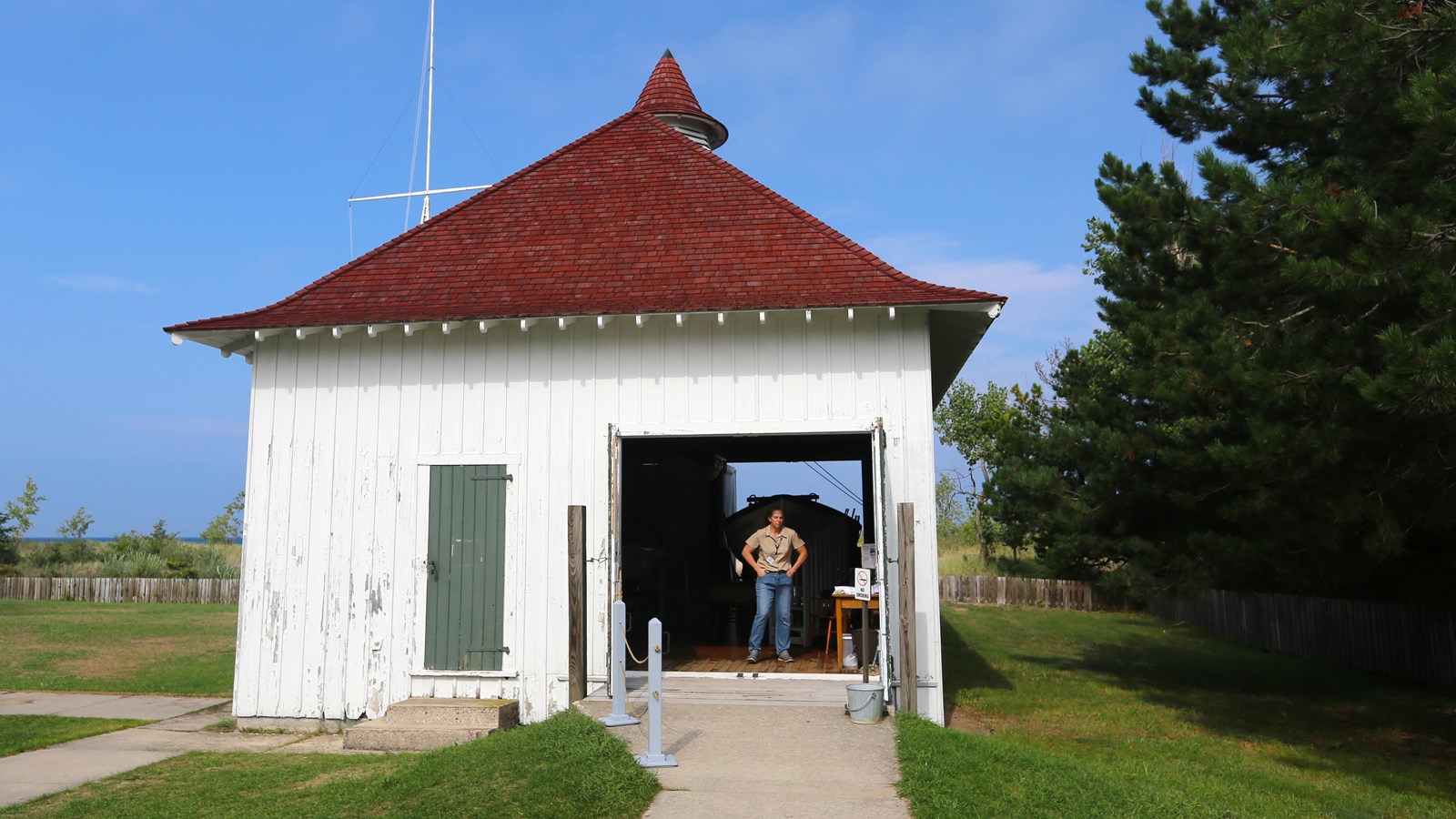 Small, white building with a red-shingled hip roof with a cupola and a volunteer standing in doorway