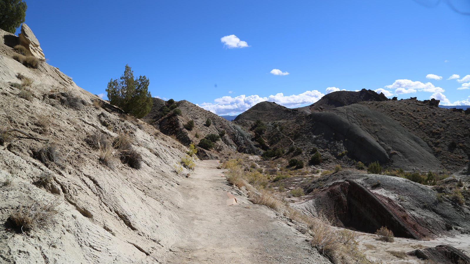 Large sandstone rock faces, adjacent to a dirt hiking trail, tilted upward with rugged scenery.