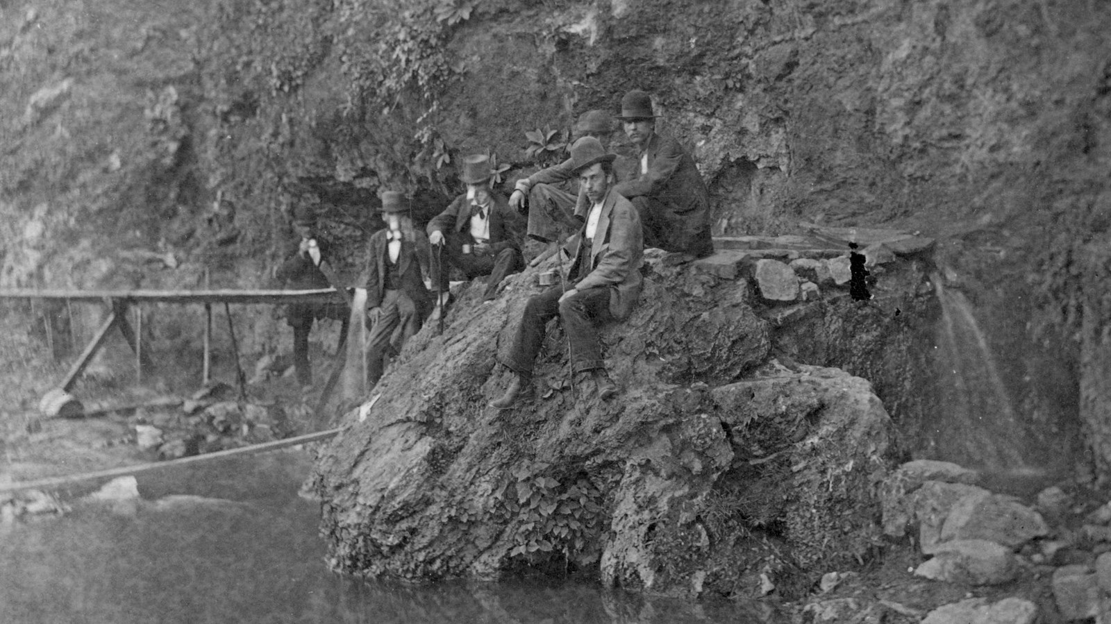 Black and White photo of six men sitting on a rock outcrop beneath a bluff along a creek.