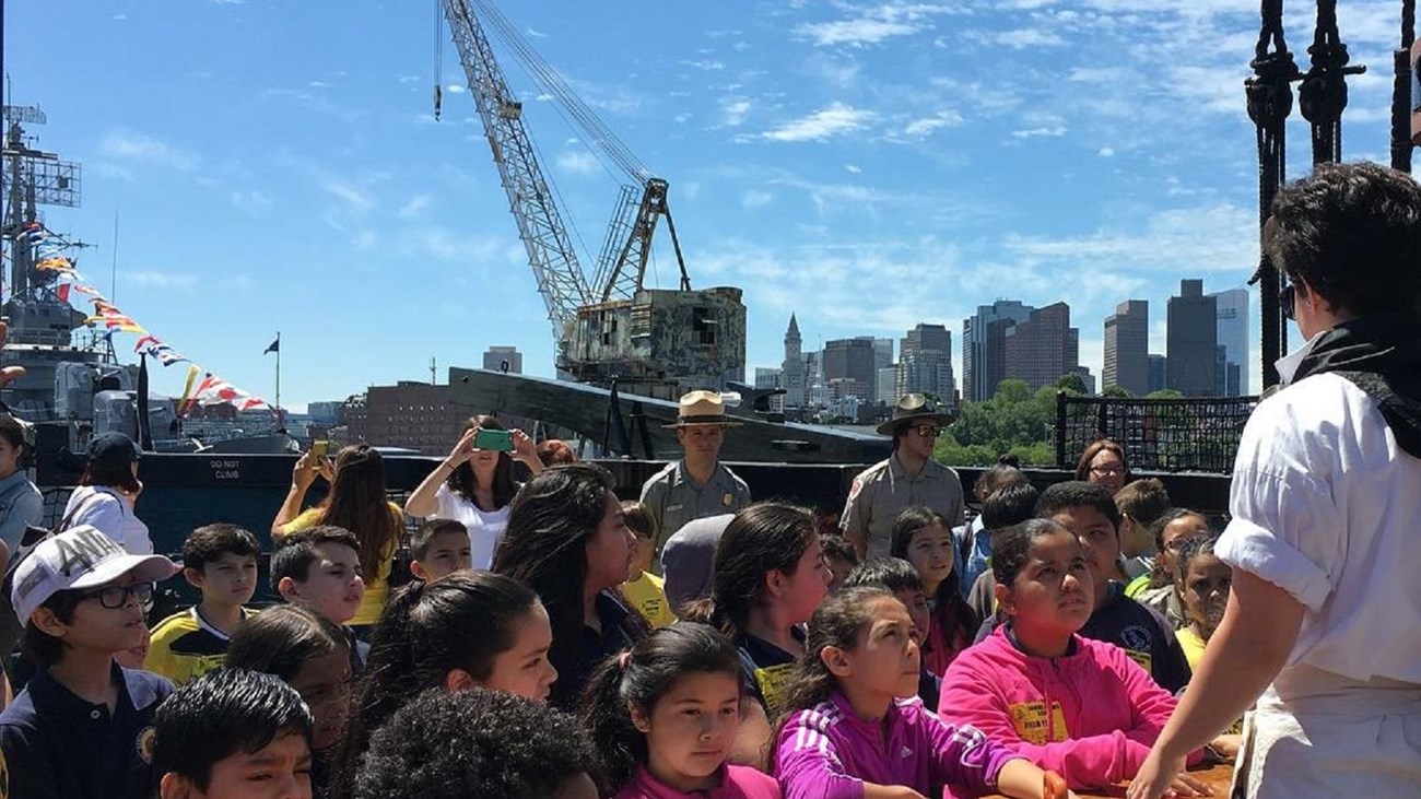 Students aboard the USS Constitution gathered around a sailor who is speaking to them.