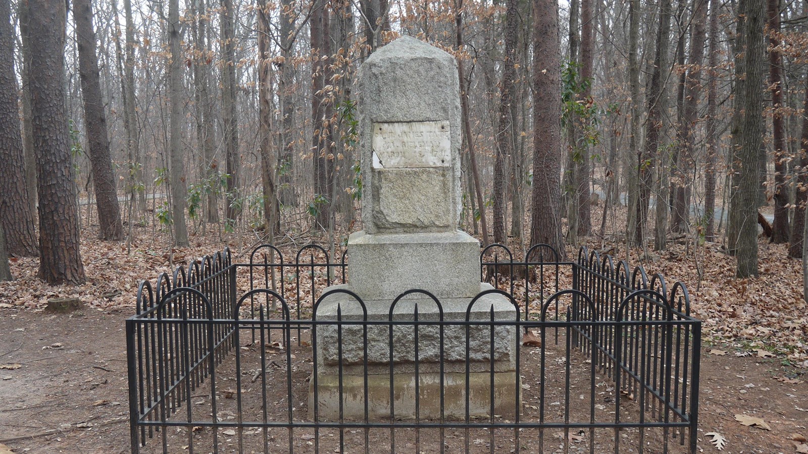 A small stone monument sits amongst trees, it has a twelve inch tall black iron fence at its base.