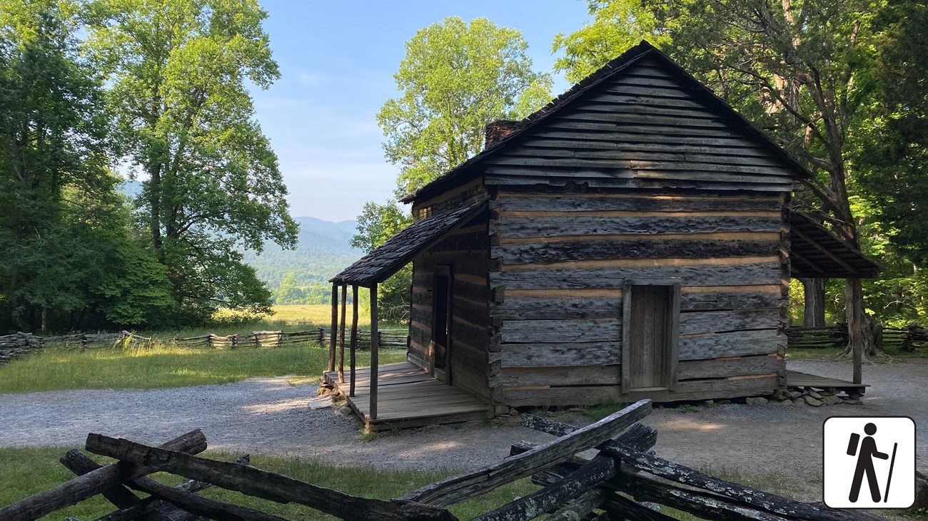 A wooden cabin with a covered porch on either side near a fence and trees. Hiker icon in corner.