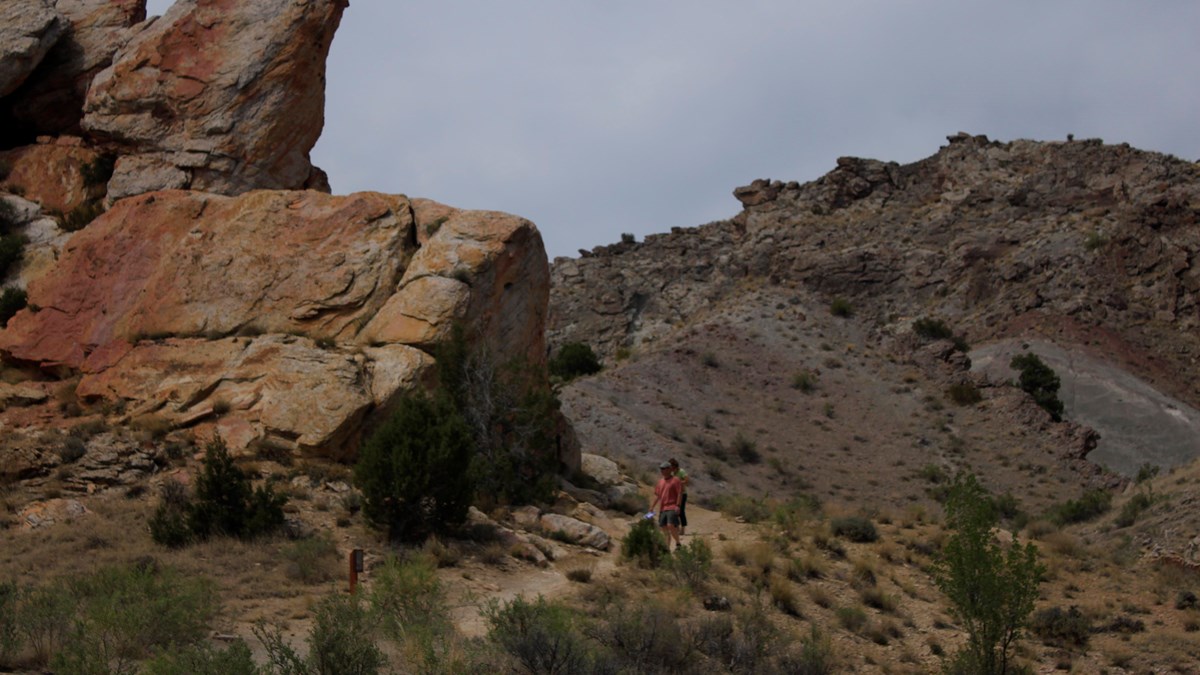 A person in a red shirt walks on a narrow trail through large colorful rock layers 