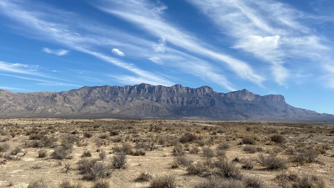 A desrt mountain range looms over a low desert foreground with sand dunes in the distance