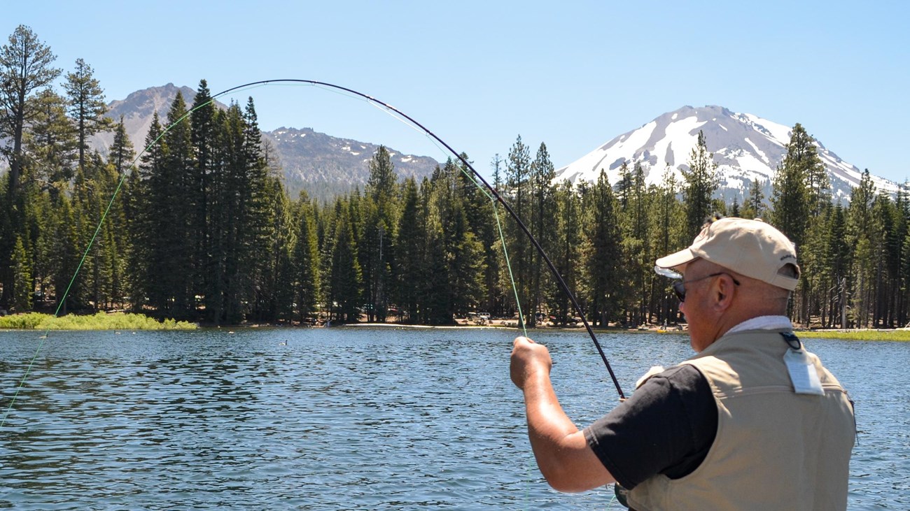 A man holds a fly fishing on a blue lake backed by conifer trees and volcanic peaks.