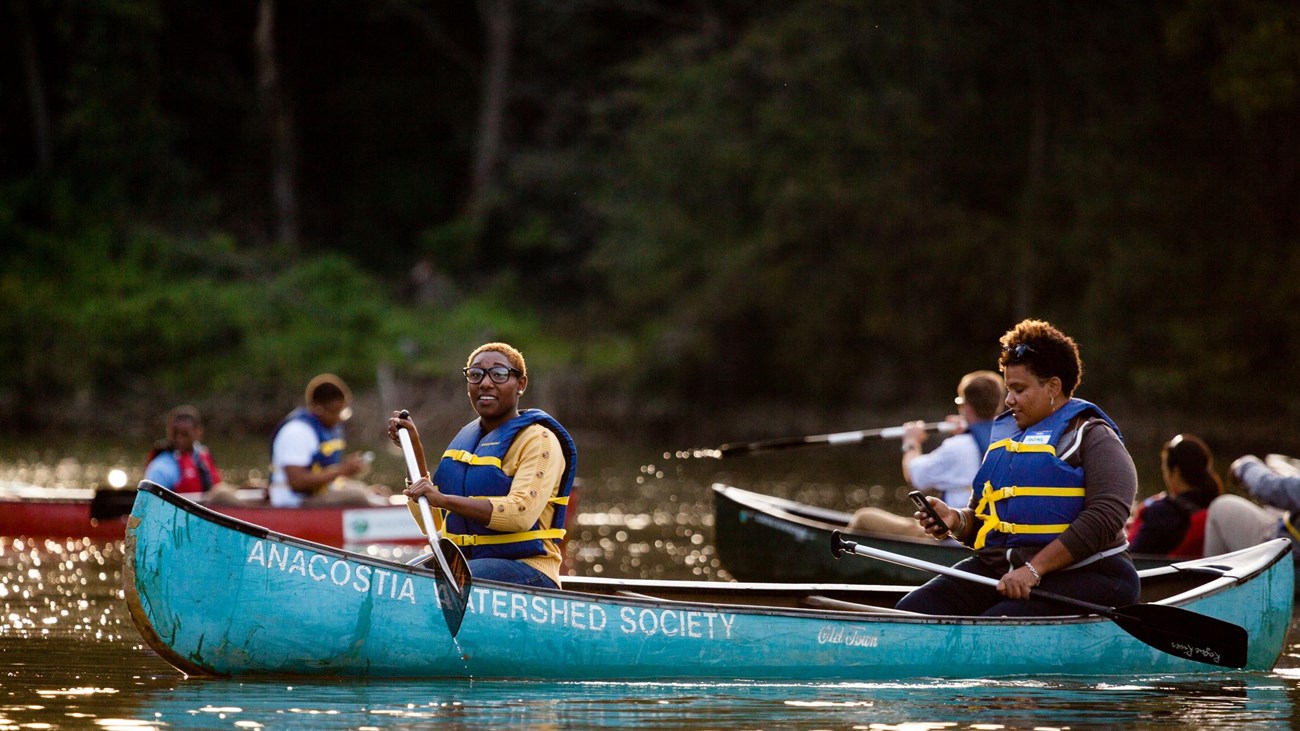 Two people paddle a canoe with two other canoes in the background. 