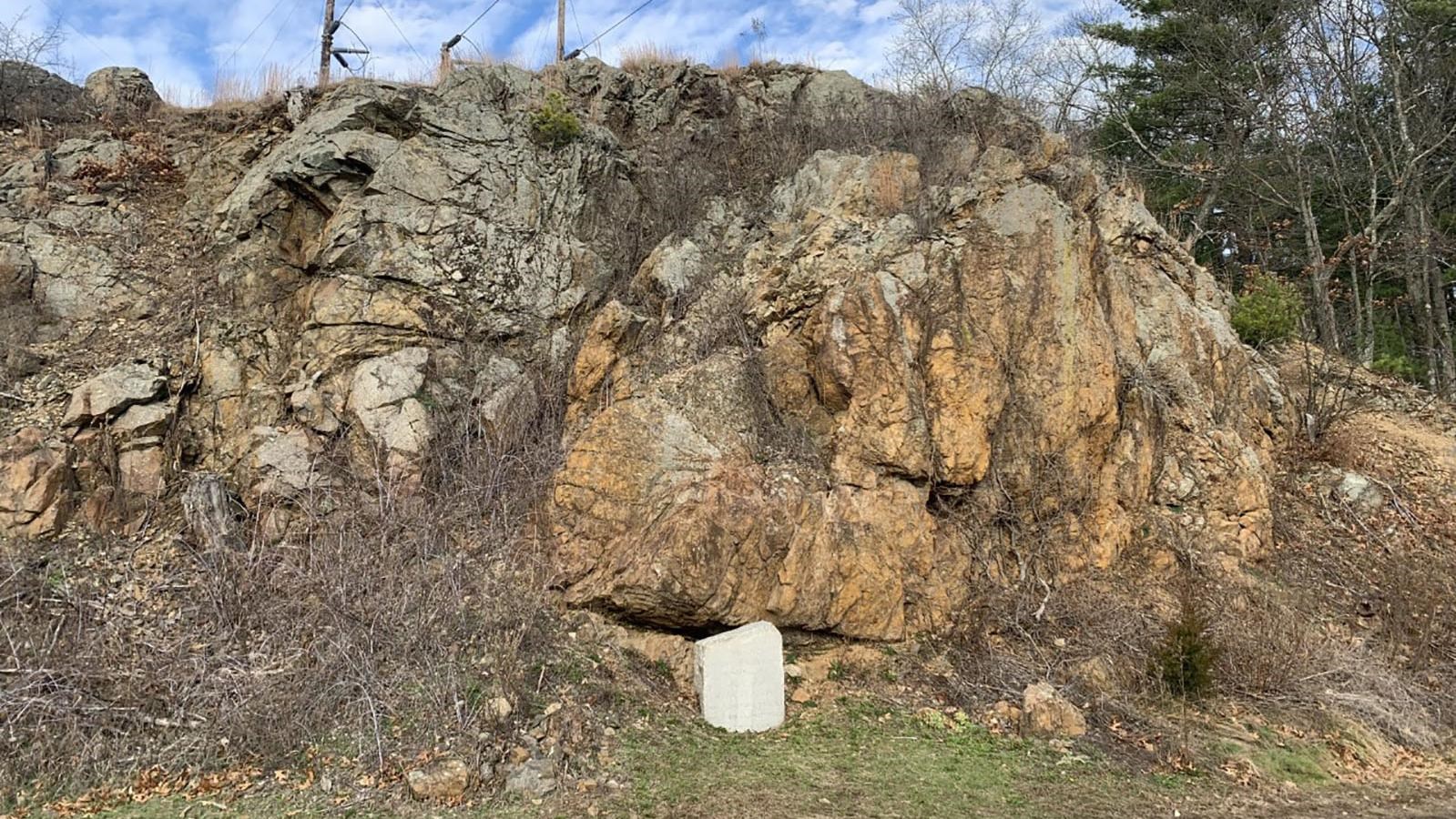 A 20 foot tall outcrop of red/gray rocks sticks out of a hill and towers over a monument and plants