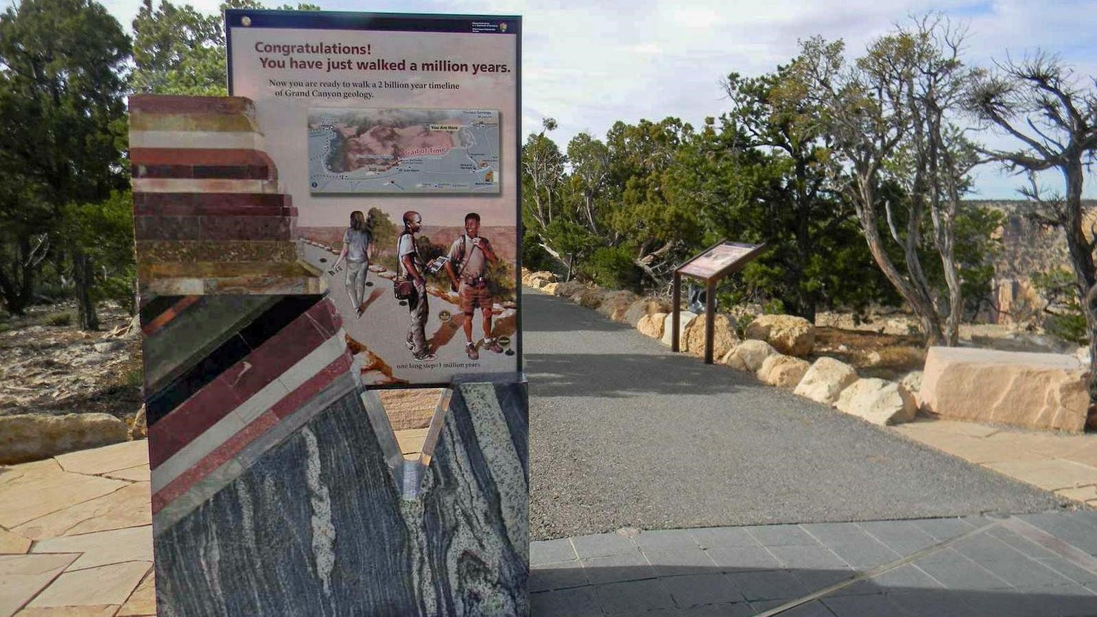 By a paved footpath, a stone monolith portal sign displays colorful geologic rock layers.