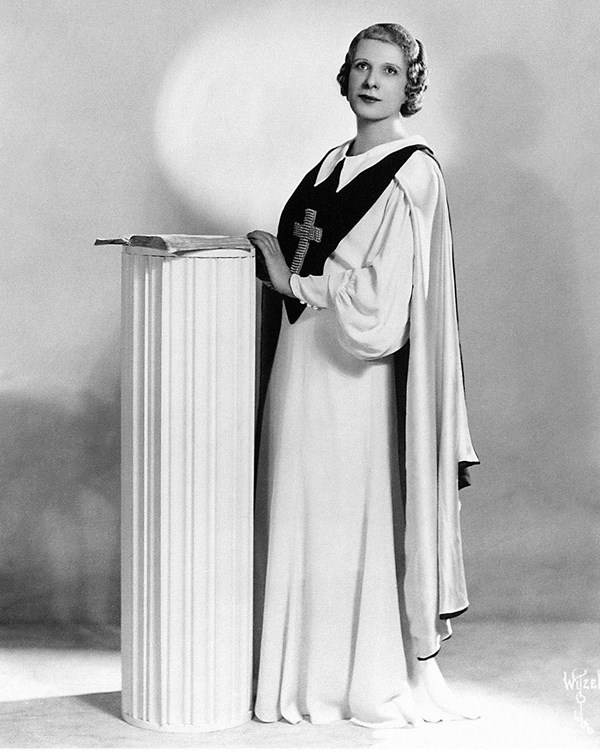 Black and white photograph of Aimee Semple McPherson standing behind a white podium.
