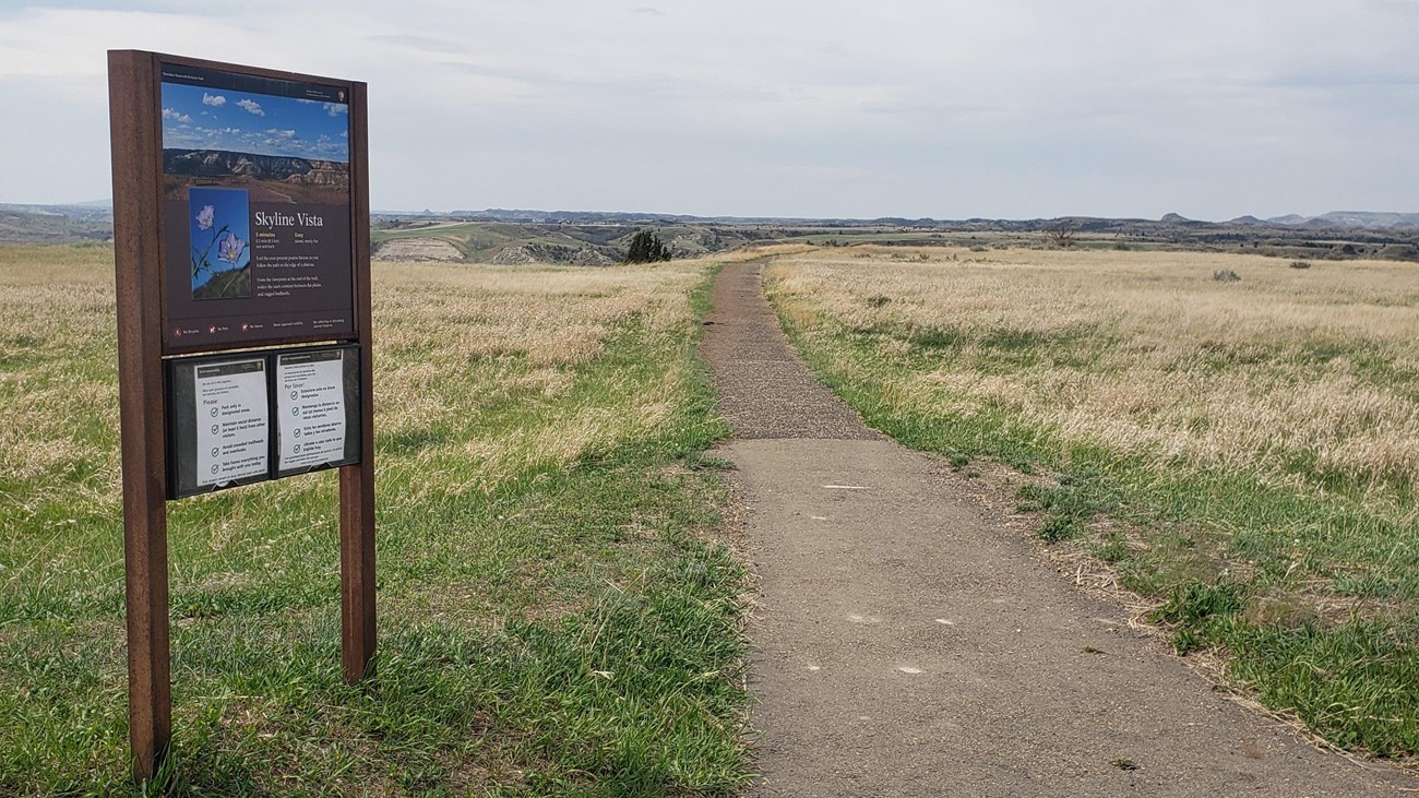 A trailhead sign next to a paved trail in a grassy area. Buttes are visible on the horizon. 