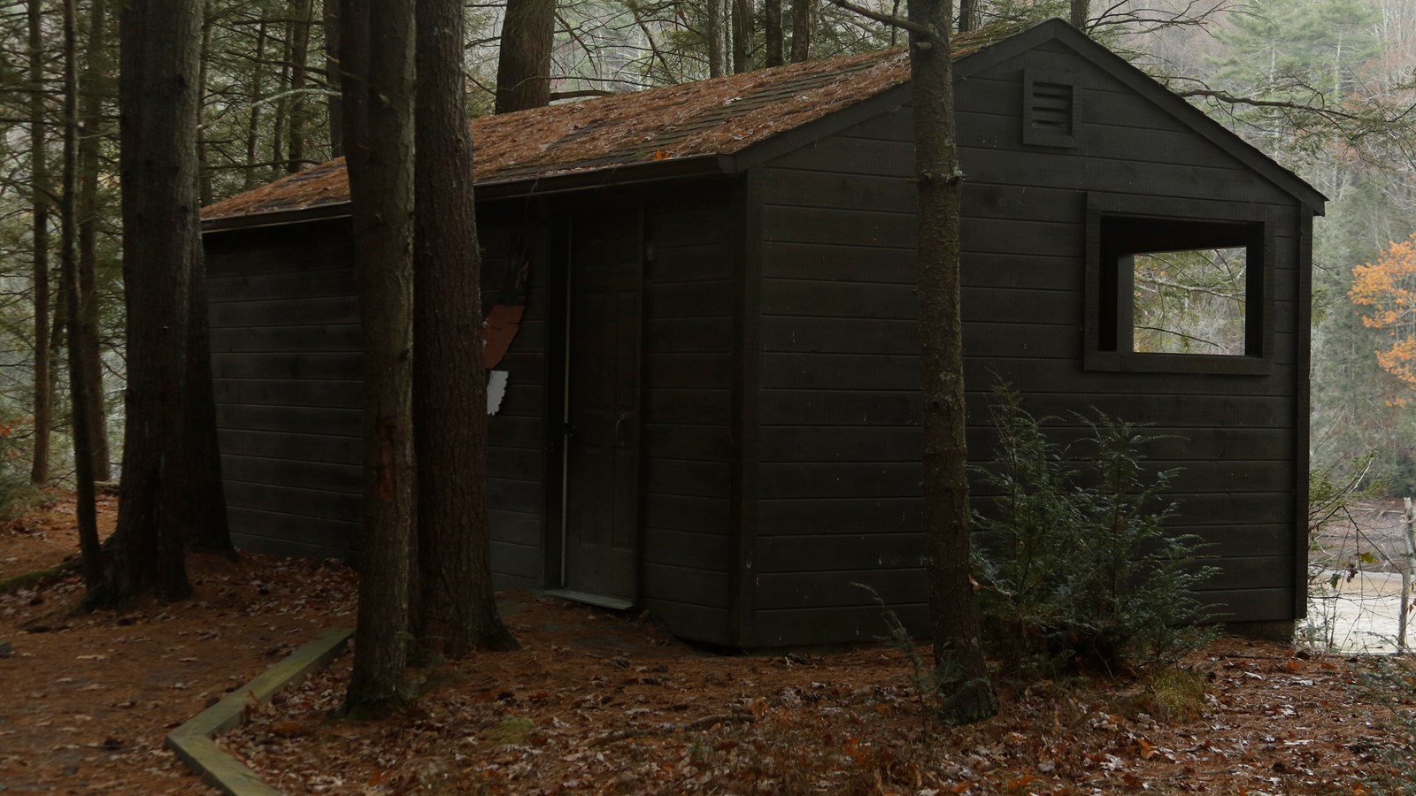 Small wooden building in a forested area. Simple path leads to the door.