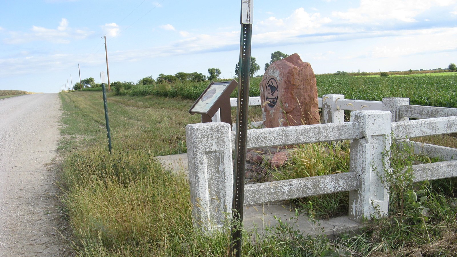 A pink stone marker sits within a white picket fence next to a dirt road, in a grassy prairie.