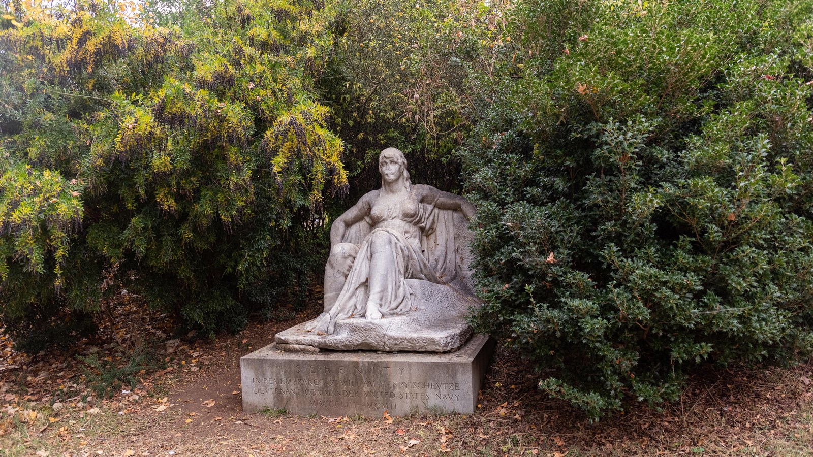A marble statue of a woman sitting in a chair. The statue is surrounded by trees. 