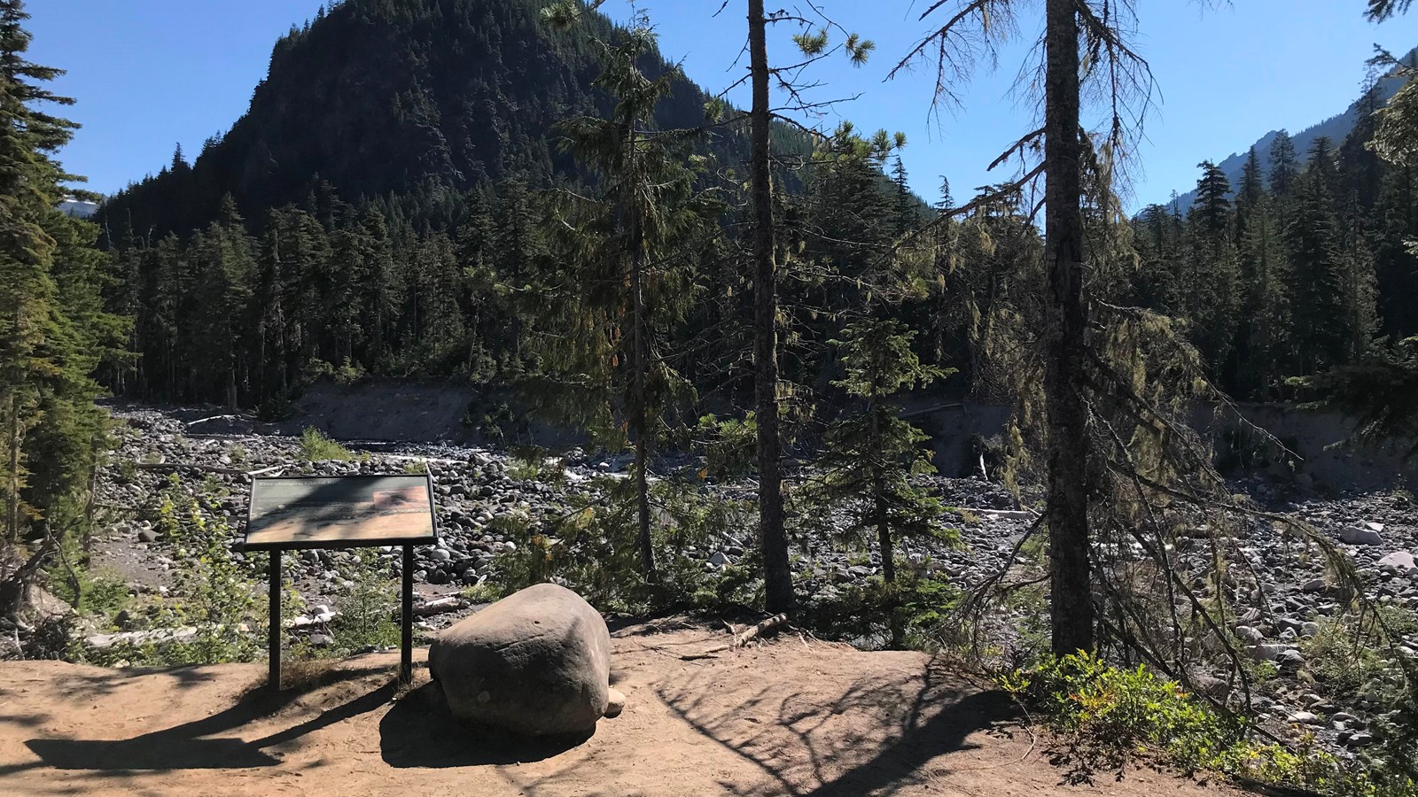 A wayside panel in a metal frame overlooks a wide, rocky riverbed and forested hills.. 