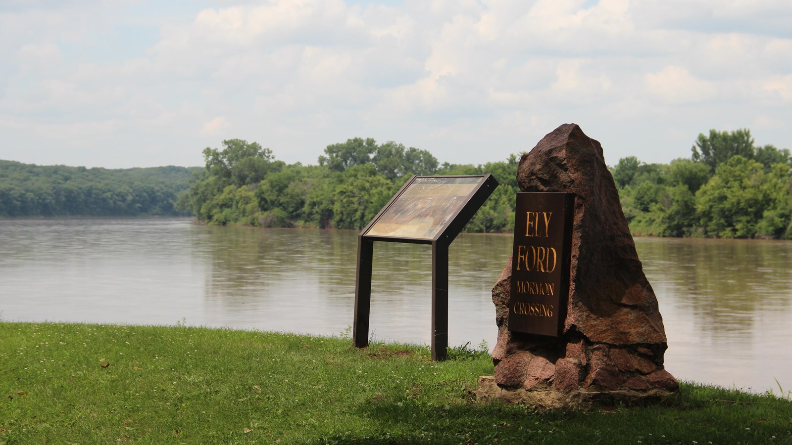 Stone monument and wayside exhibit stand on grassy riverbank overlooking cure in De Moines River.
